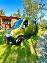 This olive-green RV rental, a converted van, sits on a sunny lawn beside a modern home with mountain views. Ideal for adventure travel.