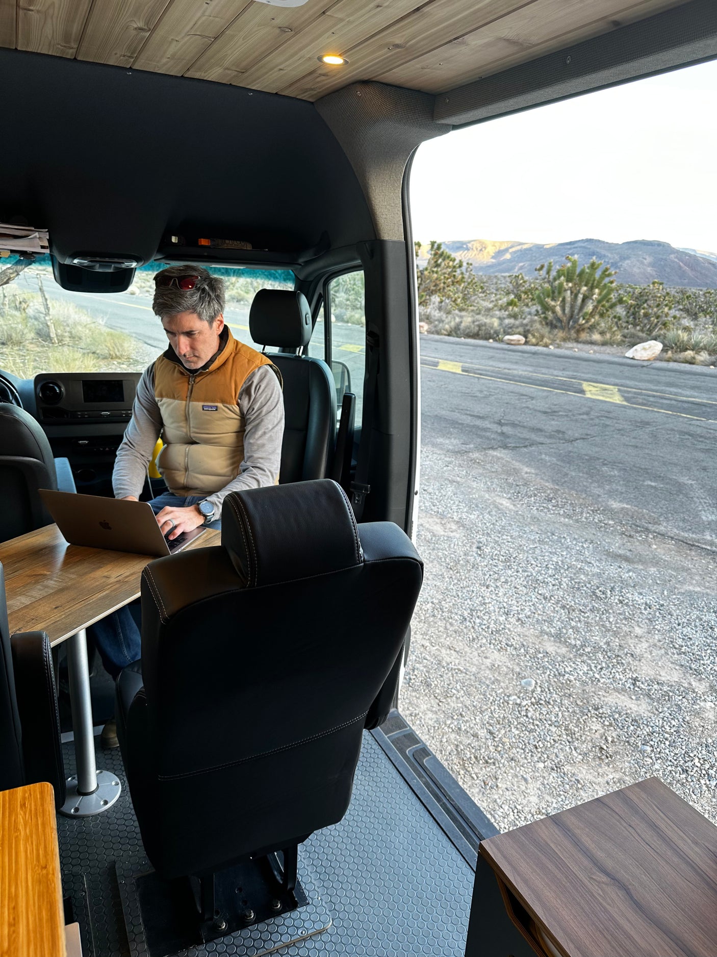 A man on a laptop in an RV rental's spacious interior. Outside, a desert road, mountains, and Joshua trees.