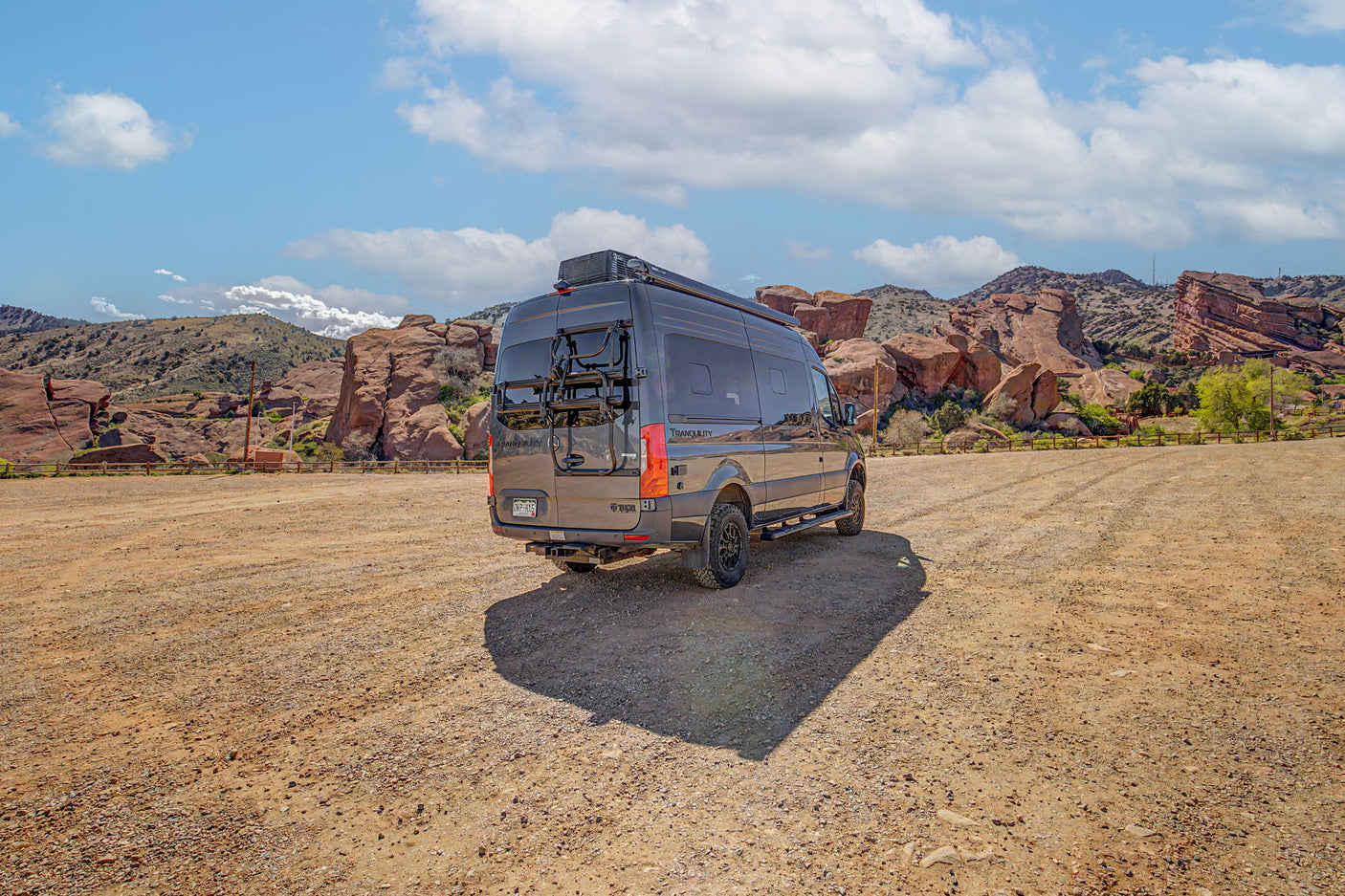Dark RV rental camper van in a gravel lot, with large red rock formations and a blue sky in the background. Adventure awaits.