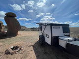 A white and black TigerMoth teardrop RV rental is parked in a dry, open landscape next to a large balanced rock formation and a fire pit under a blue sky.