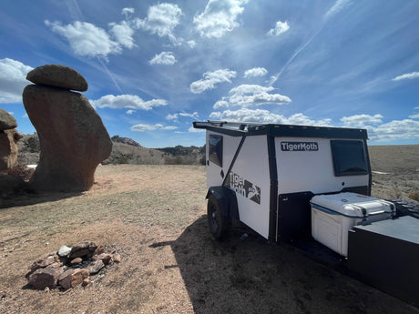 A white and black TigerMoth teardrop RV rental is parked in a dry, open landscape next to a large balanced rock formation and a fire pit under a blue sky.