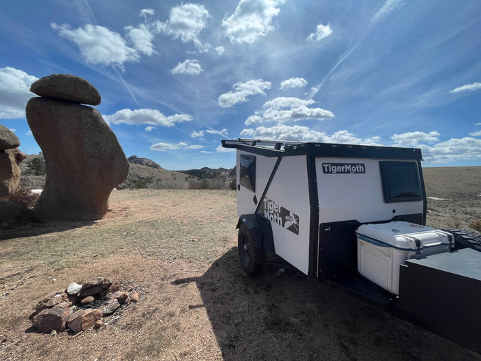 A white and black TigerMoth teardrop RV rental is parked in a dry, open landscape next to a large balanced rock formation and a fire pit under a blue sky.