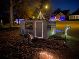 A small, holiday-decorated RV rental camper sits on a lawn with festive lights. Nearby homes also sparkle brightly at night.