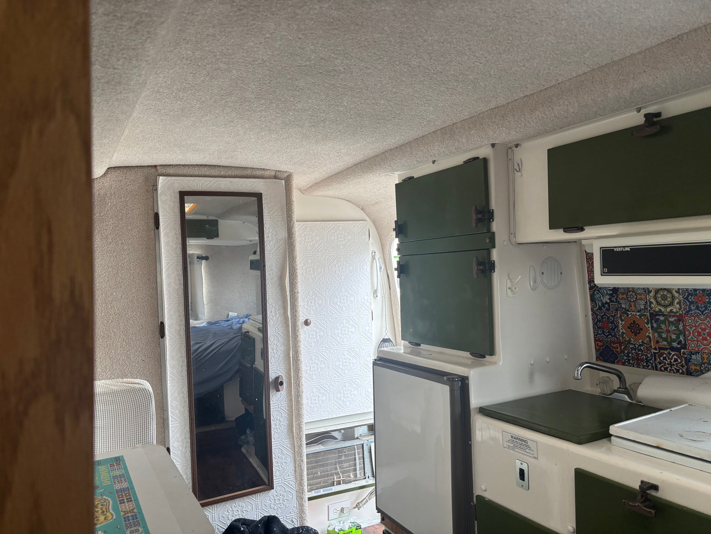 RV rental interior: a compact kitchenette with green cabinets, sink, fridge, and a mirrored door reflecting the sleeping area.