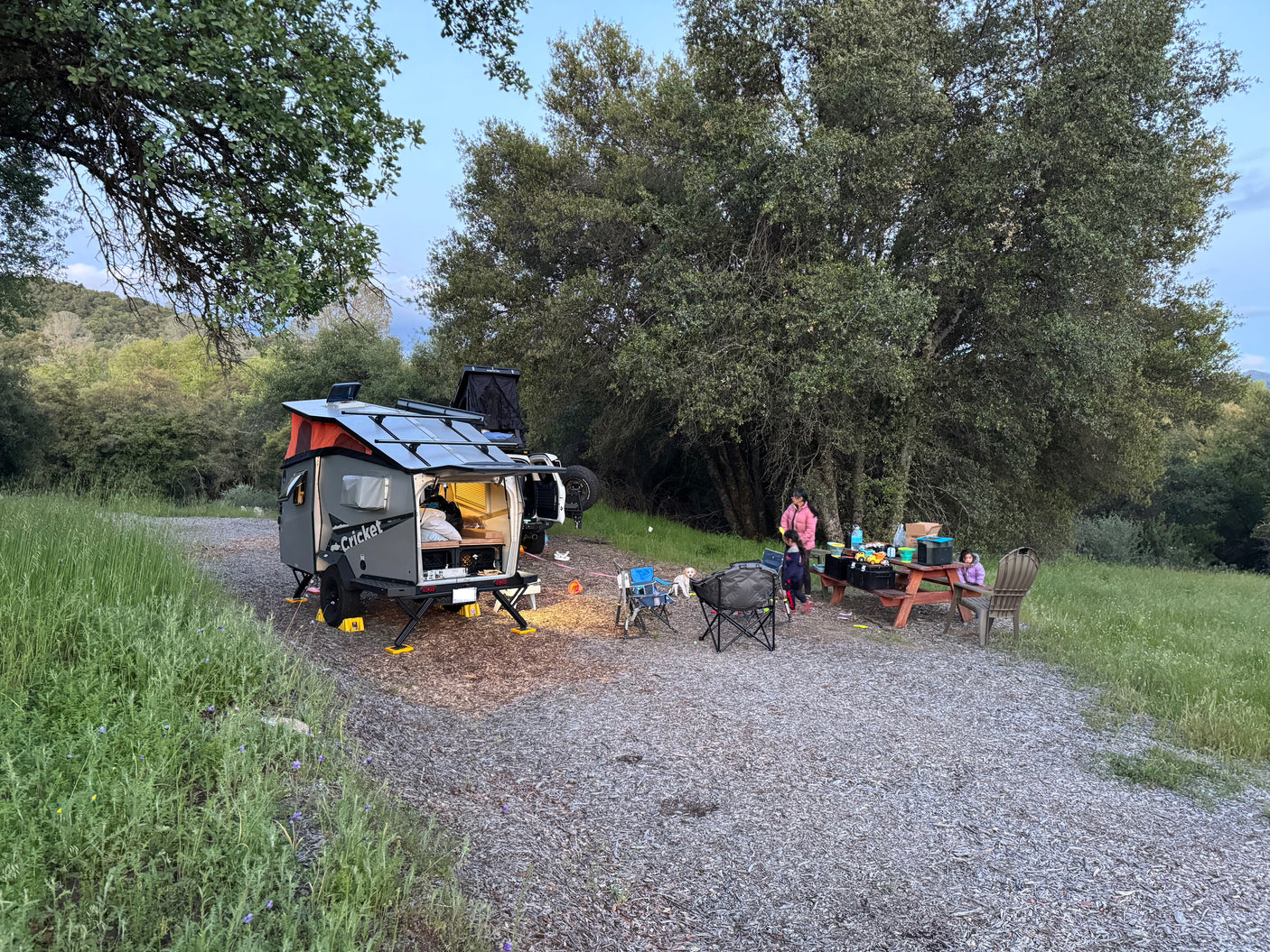 Family enjoying their rv rental 'Cricket' trailer and SUV set up at dusk. Camp chairs, picnic table, and dog visible.