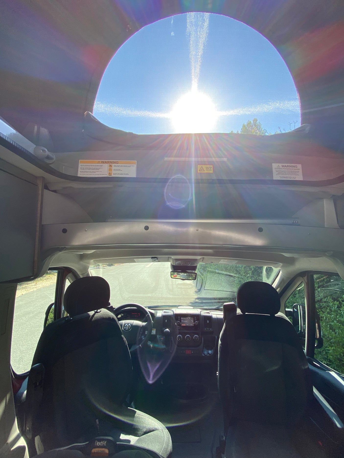 Bright sun streams through an RV rental's rooftop opening, illuminating the driver's cockpit, road ahead, and a NextFit child car seat.