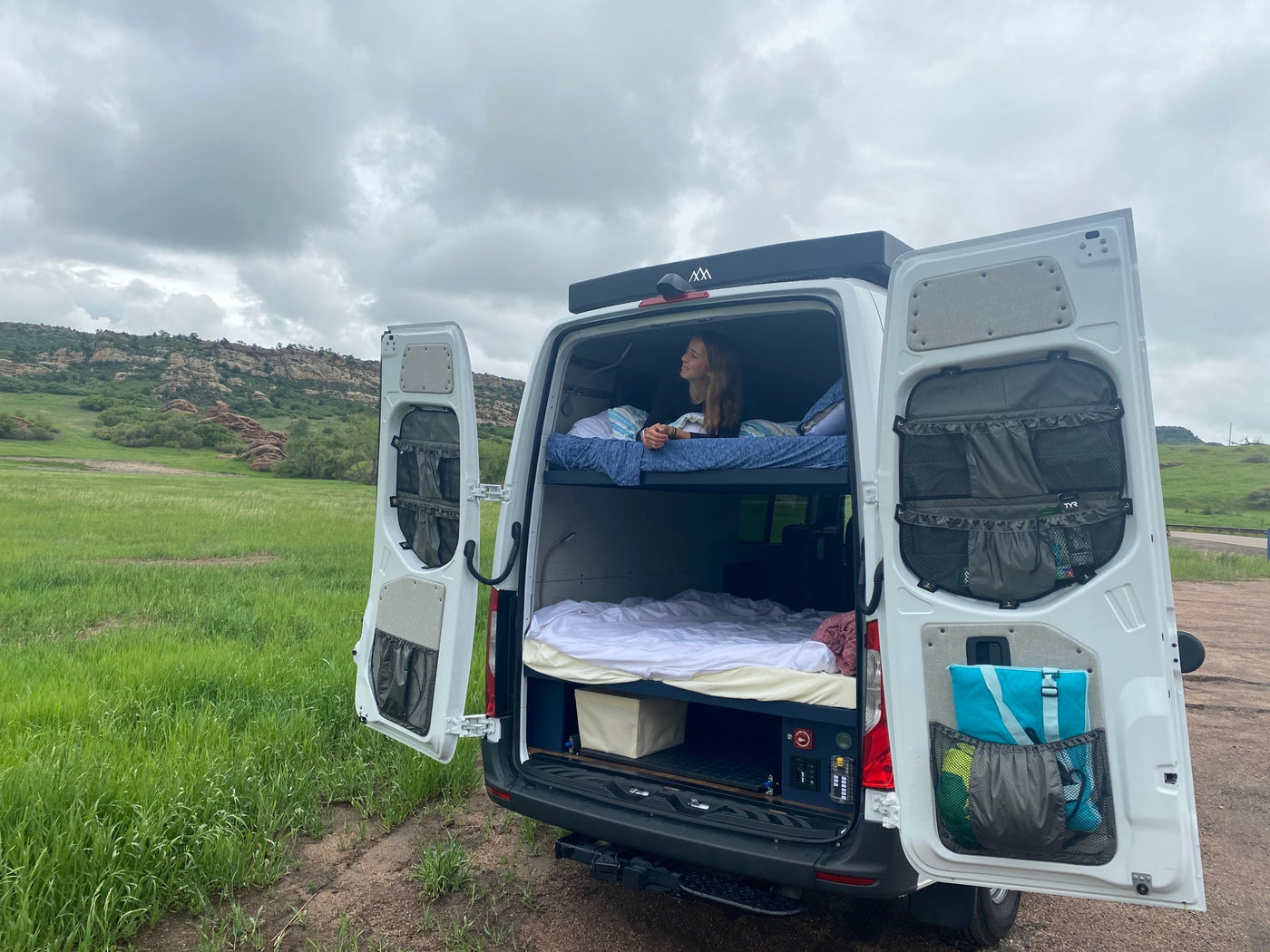 Relaxing in an RV rental van, a woman on the bunk bed enjoys views of a green field and rocky mountains. Perfect for van life. — photo 8