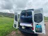 Relaxing in an RV rental van, a woman on the bunk bed enjoys views of a green field and rocky mountains. Perfect for van life.