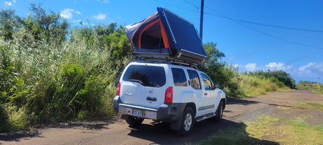 A white Nissan Xterra SUV with a black and orange roof-top tent deployed, parked on a dirt road. An ideal RV rental for outdoor camping.