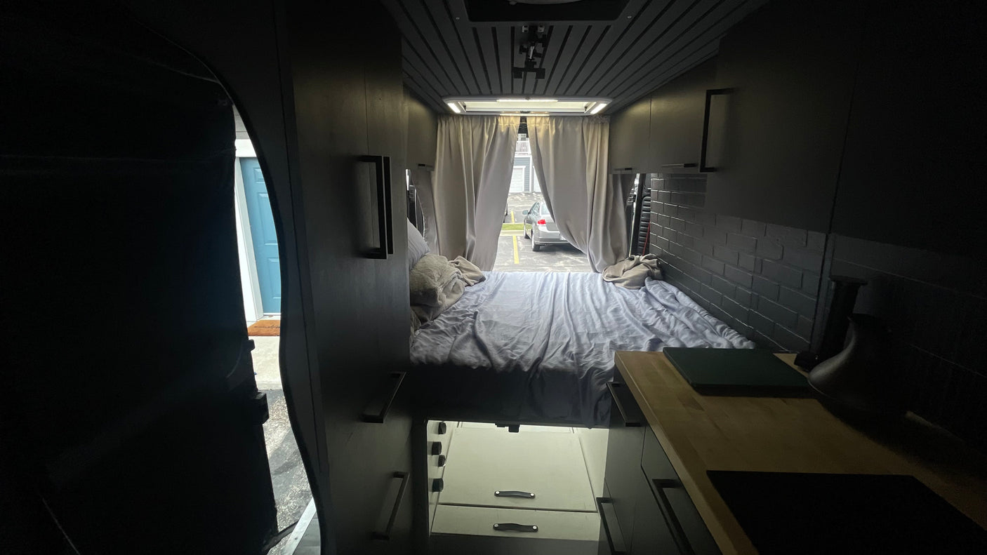 Interior of a modern RV rental featuring a comfortable bed, sleek storage, and a kitchen counter with an outdoor view.