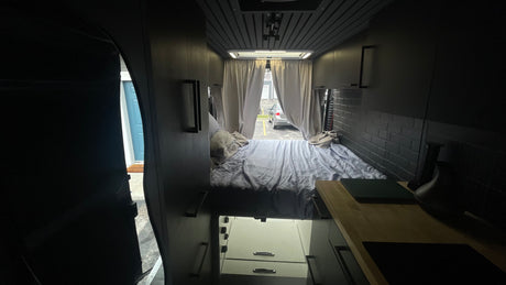 Interior of a modern RV rental featuring a comfortable bed, sleek storage, and a kitchen counter with an outdoor view.