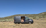 Woman relaxes in an **rv rental** camper van in a dry, shrubby landscape under clear skies, perfect for off-grid travel.
