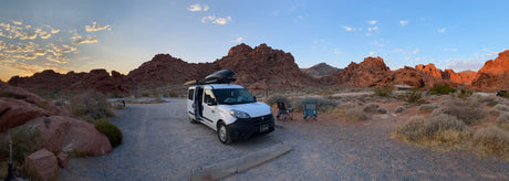 An RV rental van and camping chairs rest among stunning red rock formations in a scenic desert landscape at sunset.