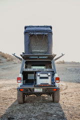 Rear view of a white Jeep Gladiator with a deployed rooftop tent and an open truck camper revealing a fridge. Great for RV rental expeditions!