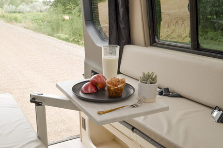 A cozy RV rental setup with breakfast (milk, apples, muffin) on a swing-out table, overlooking a scenic road.