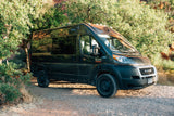 Experience nature with this black Ram ProMaster rv rental camper van, parked on a sunlit gravel road amidst green trees.