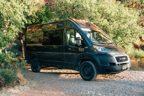 Experience nature with this black Ram ProMaster rv rental camper van, parked on a sunlit gravel road amidst green trees.