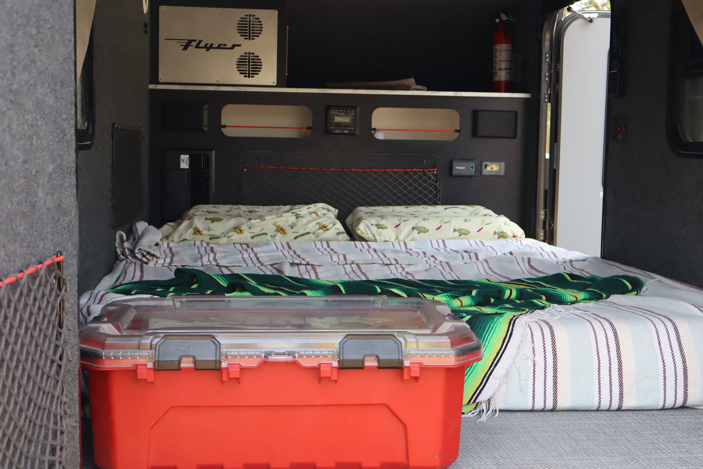 Detailed view of an RV rental sleeping space with patterned bedding, overhead controls, and a red storage toolbox.
