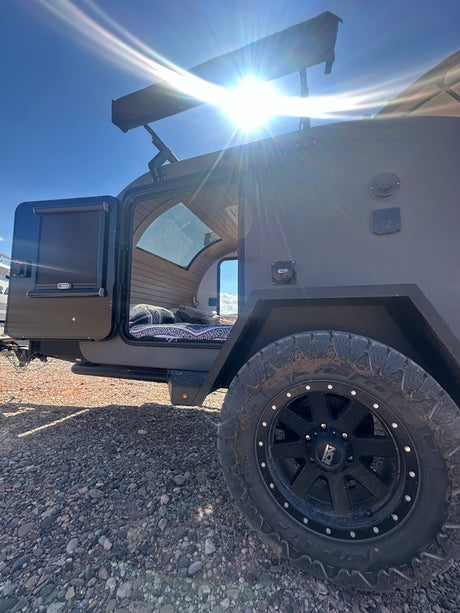 Adventure-ready RV rental trailer on gravel. Open door reveals a cozy, wood-paneled interior for off-road camping.