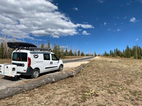 White RV rental van with roof cargo & cooler on a scenic road. Bright blue sky, clouds, and forest with fallen log.