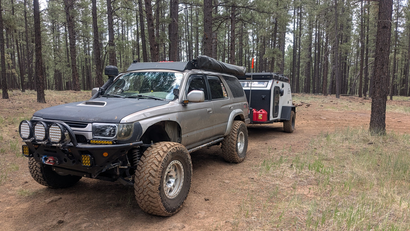 Rugged off-road SUV towing a compact camper, ideal for an RV rental camping adventure in a dense pine forest. Overlanding setup.