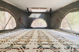 Inside an RV rental: a spacious pop-top bed with a Southwestern blanket and mesh windows overlooking a forest campsite.