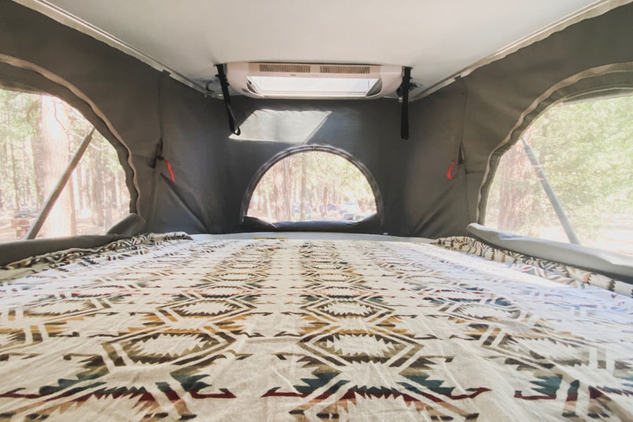 Inside an RV rental: a spacious pop-top bed with a Southwestern blanket and mesh windows overlooking a forest campsite.