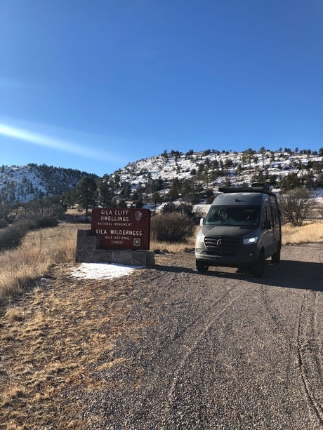 Mercedes RV rental van at Gila Cliff Dwellings sign, snowy Gila Wilderness mountains in background.
