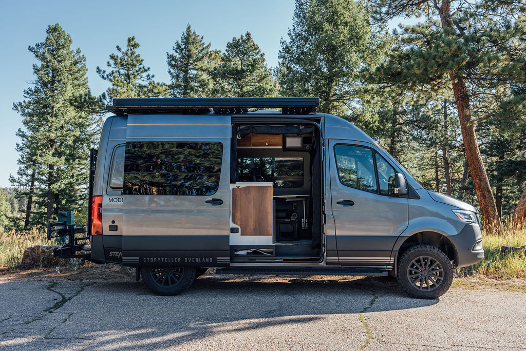  framed by yellow wildflowers and majestic mountains.|A sleek Mercedes Sprinter RV rental is parked by a serene river