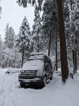 Snowy RV rental Mercedes Sprinter van parked in a winter forest campsite. Deep snow covers trees and ground.