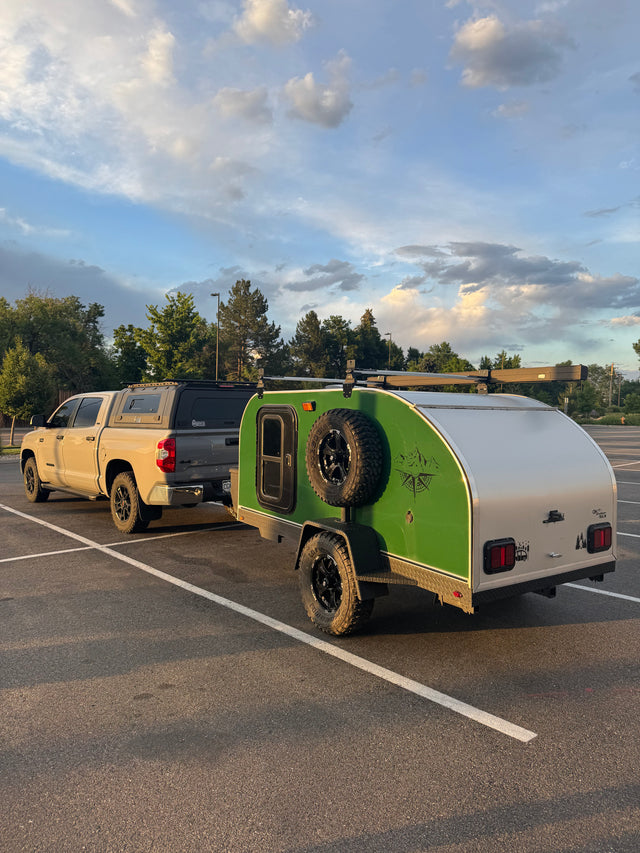 Adventure-ready RV rental: Tan pickup truck with camper shell towing a green teardrop trailer, perfect for camping adventures under a cloudy sky.