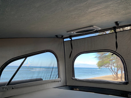 Interior of an RV rental showing two windows with scenic views of ocean waves, a sandy beach, and trees.