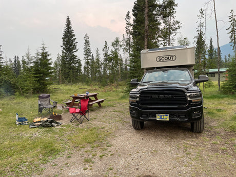  on a vast beach with ocean and mountains under cloudy skies.|Black RAM truck with a SCOUT truck camper