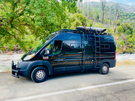 Black RV rental van beside a vibrant river, surrounded by lush mountains and trees. Equipped for adventure.