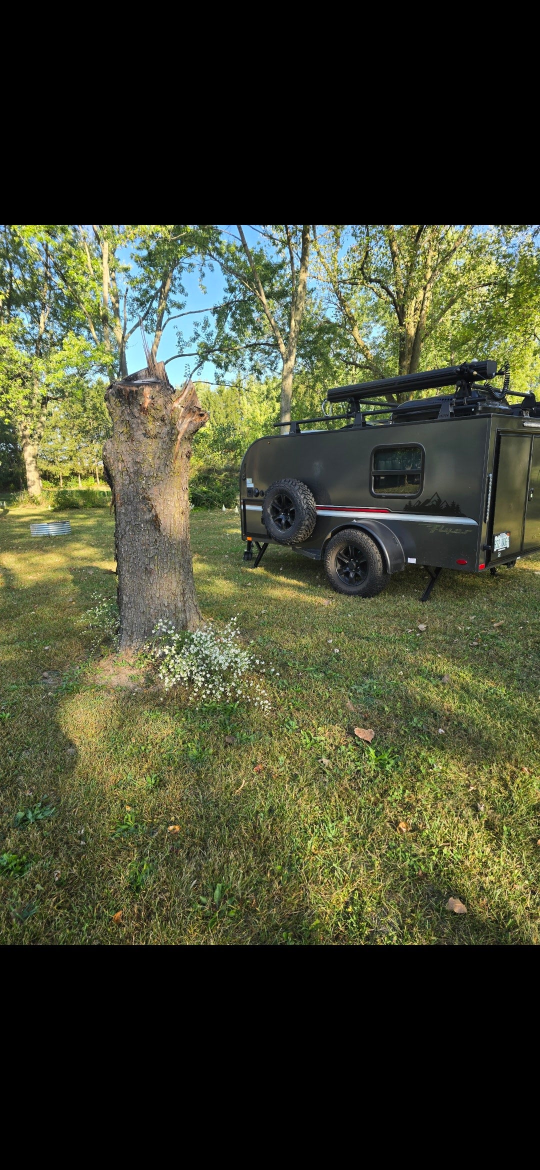 A dark grey teardrop rv rental with off-road tires and roof rack, parked on a grassy campsite near a large tree stump.