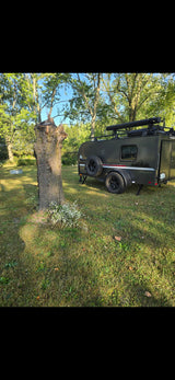 A dark grey teardrop rv rental with off-road tires and roof rack, parked on a grassy campsite near a large tree stump.