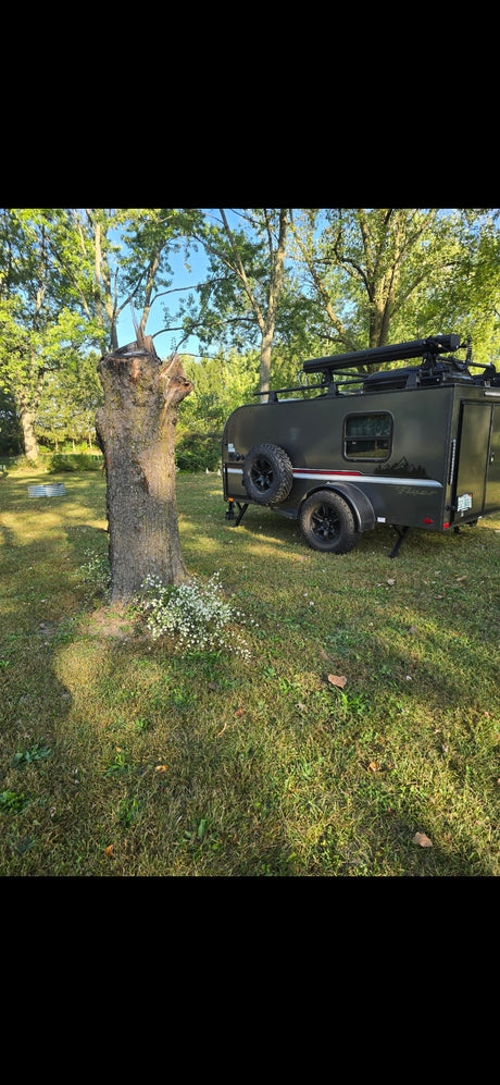 A dark grey teardrop rv rental with off-road tires and roof rack, parked on a grassy campsite near a large tree stump.