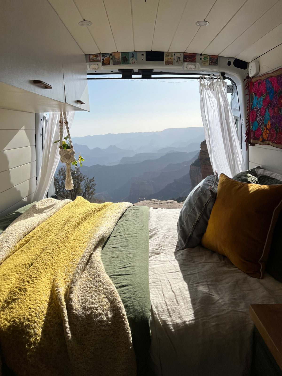 Inside a comfy RV rental, a bed with soft blankets faces an open back, revealing layers of serene mountain canyons.
