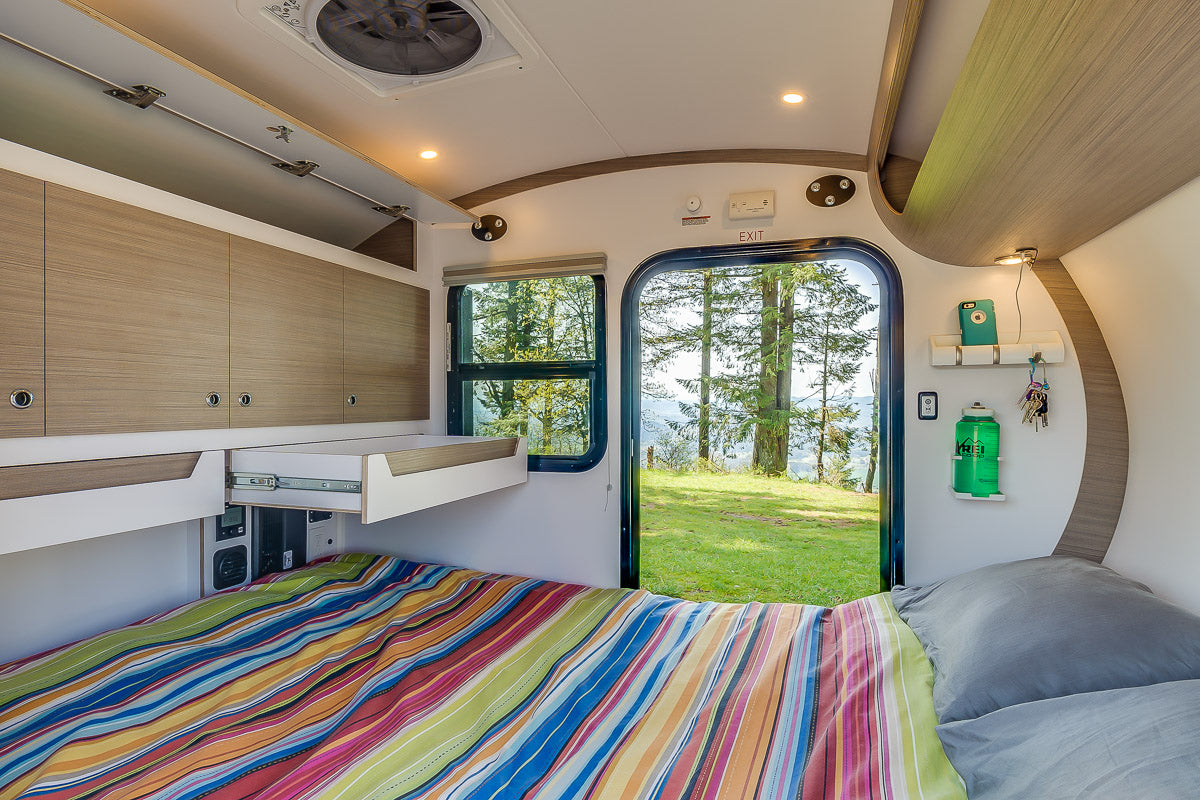 Modern RV rental interior with a colorful bed, smart storage, and a scenic view of nature through the open door and window.