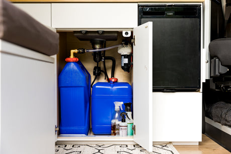 RV rental under-sink area: fresh & grey water tanks, pump, plumbing, and Dometic fridge in a camper van.