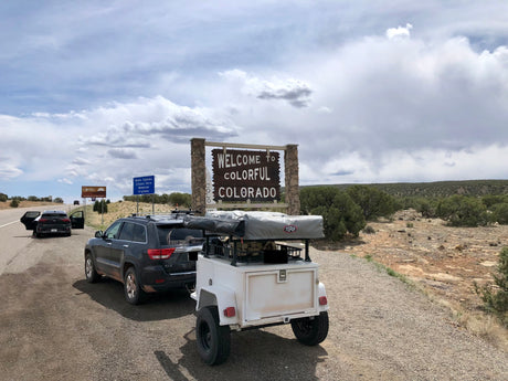 Entering Colorado! SUV with overland trailer (like an rv rental) at the state line sign, ready for a scenic road trip adventure.