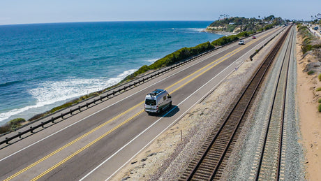 Coastal rv rental trip: White RV drives on a sunny highway next to the ocean & parallel train tracks.