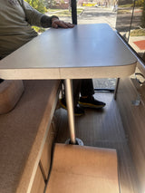Interior of an rv rental showing a person seated at a light table with a center pole, between upholstered benches on wood-look flooring.