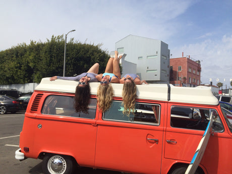 Three women in swimwear lie upside down on a bright orange VW camper van. Great for an rv rental beach adventure!