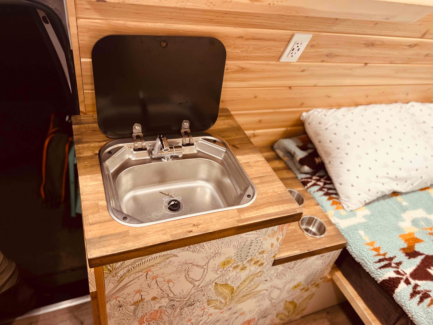 This RV rental showcases a compact sink on a wood countertop, with light wood paneling, an outlet, and a cozy bed area.