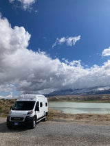A white Ram ProMaster camper van by a serene lake with snow-capped mountains under a cloudy sky. Perfect for an RV rental adventure.