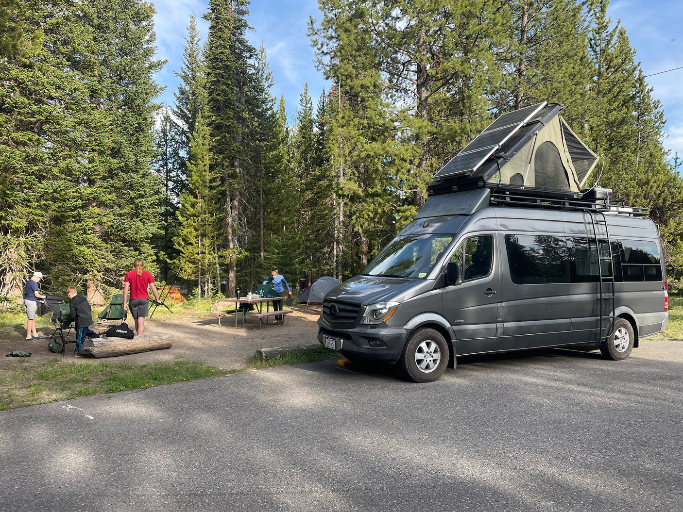 A grey RV rental van with solar panels & a rooftop tent is parked at a forested campsite where people are setting up.