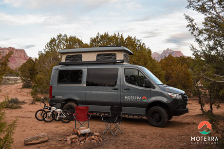 Moterra El Capitan RV rental campervan parked for camping, with bikes and chairs by a campfire in a desert landscape.