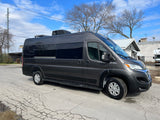 Dark gray RV rental camper van with tinted windows and roof AC. Parked on a cracked asphalt lot, ready for travel.
