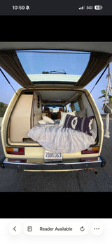 Inside a cozy vintage camper van RV rental, the open tailgate reveals a bed with patterned pillows, storage, and a hanging plant.